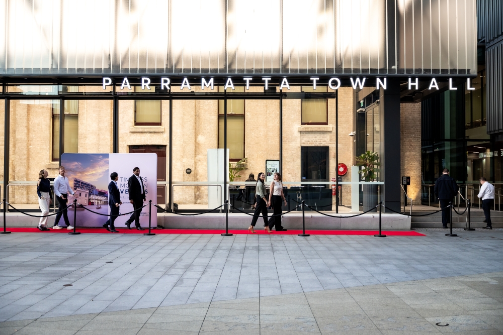 People walking on a red carpet in front of Parramatta Town Hall, with a backdrop of banners and an elegant modern facade. Event atmosphere.