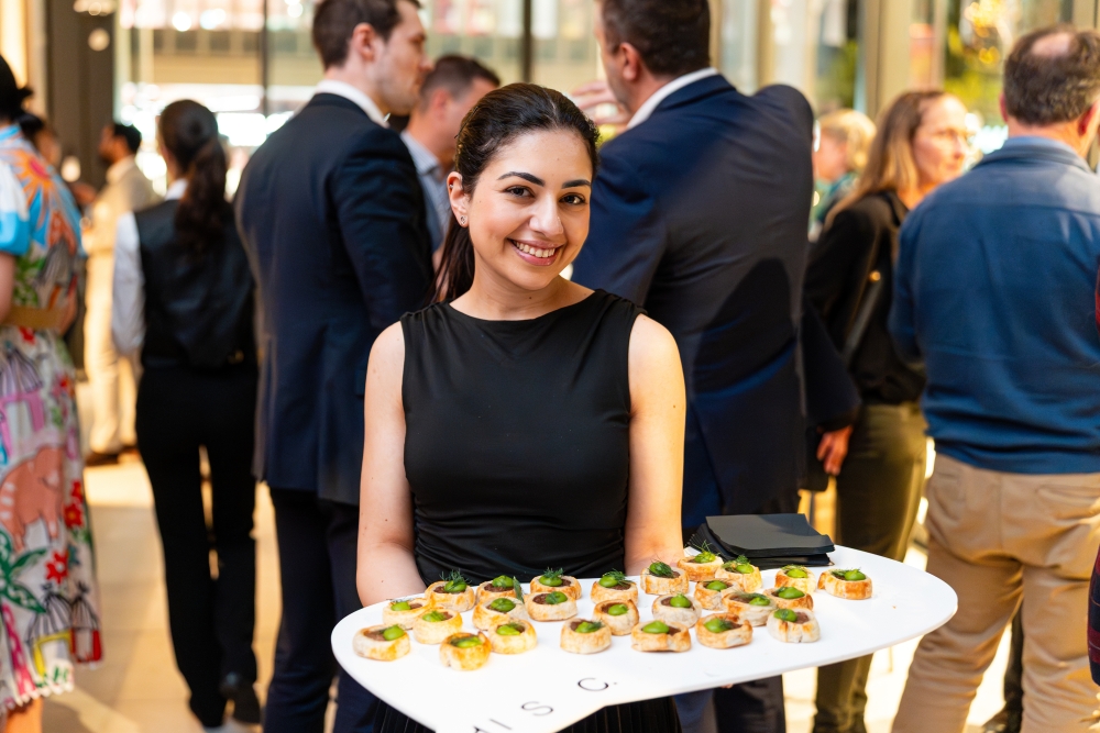 Smiling woman in black dress holding a tray of appetizers at a social event. People in stylish attire converse in the background, creating a lively atmosphere.