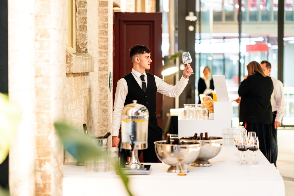 A waiter in a black vest inspects a wine glass at a modern event. Behind him, guests mingle near a bar. The setting is upscale and elegant.
