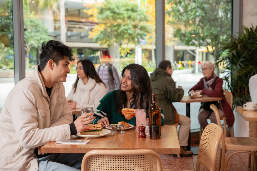 A couple enjoys lunch at a cafe, sharing smiles and conversation. The warm setting is lively, with other patrons in the background and greenery outside.