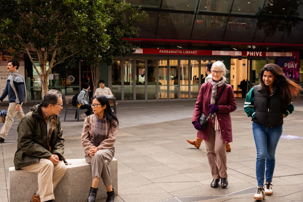 Two people sit on a concrete bench conversing under a tree in an urban plaza. Nearby, two women walk together, smiling. Parramatta Library is in the background.