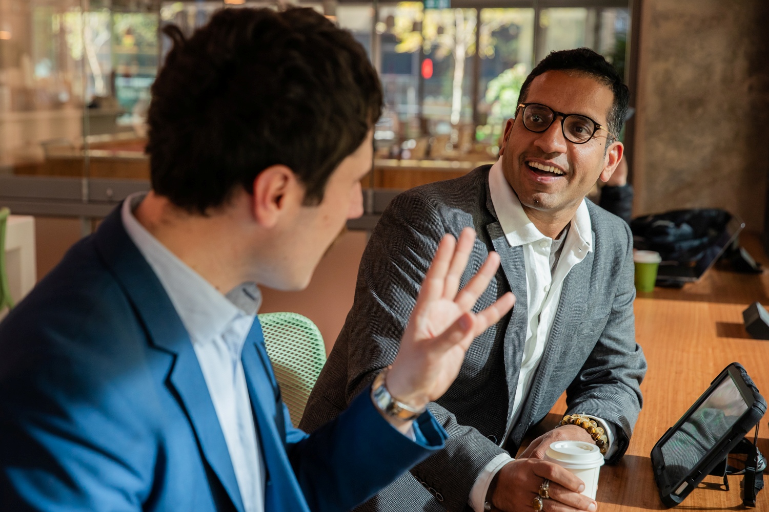 Two men in a casual business setting, smiling and conversing. One holds a coffee cup, the other gestures animatedly. A tablet rests on the table.