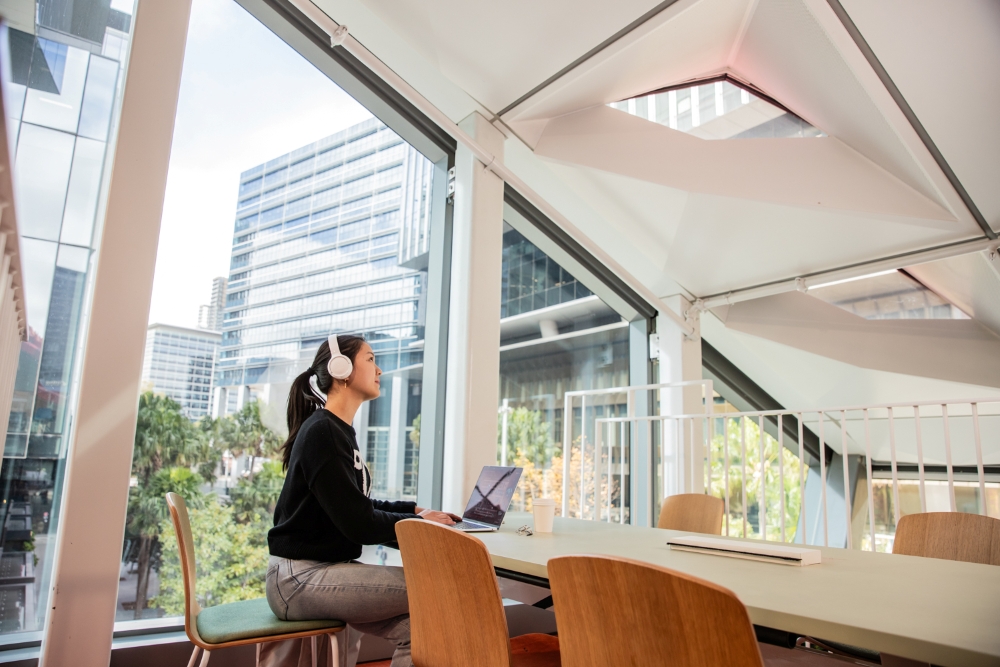 A woman wearing headphones sits at a table with a laptop in a modern, bright office with large windows. She appears relaxed and focused, surrounded by cityscape views.