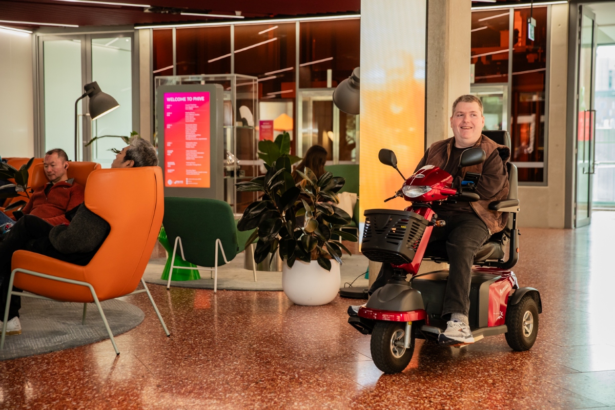 A man on a red mobility scooter smiles in a modern lobby with orange chairs, plants, and a digital screen. The atmosphere is welcoming and inclusive.