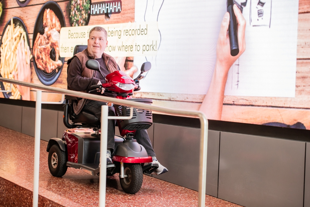 A man on a red mobility scooter smiles near a large digital screen displaying food images and text. The scene feels lively and engaging.