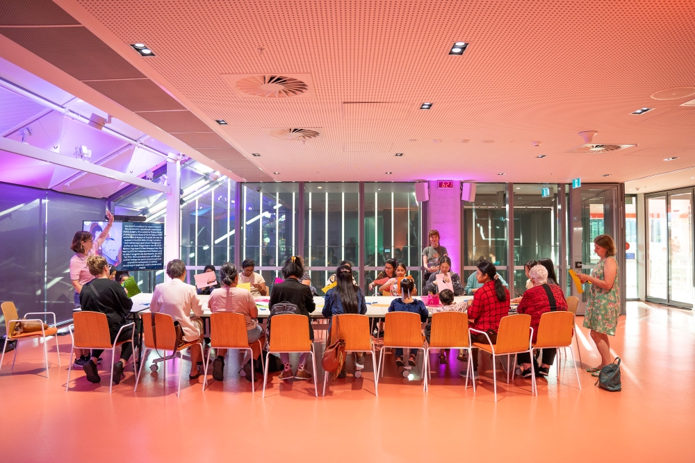 A group of people engage in a workshop in a brightly lit room with modern decor and large windows. The atmosphere is collaborative and focused.