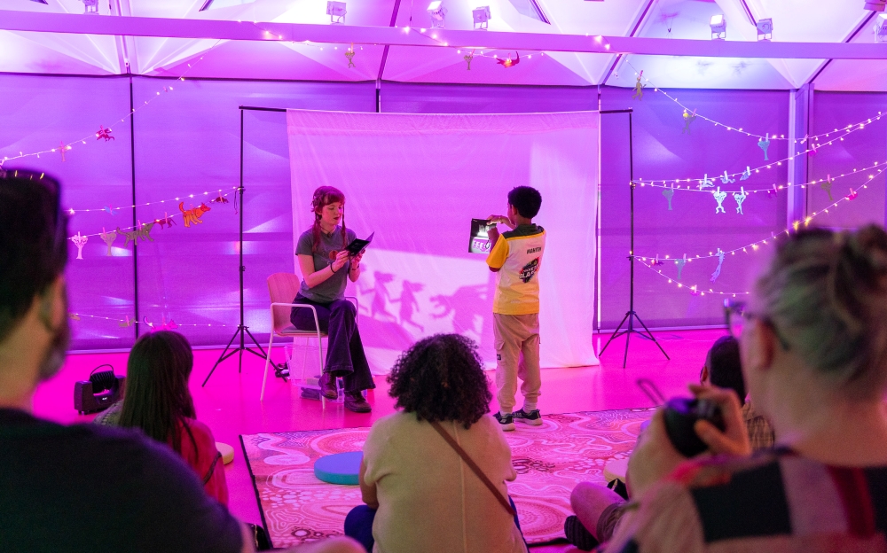 A woman reads to a child holding a tablet in a colorful room. Shadow puppets are projected on a screen. The atmosphere is warm and creative.