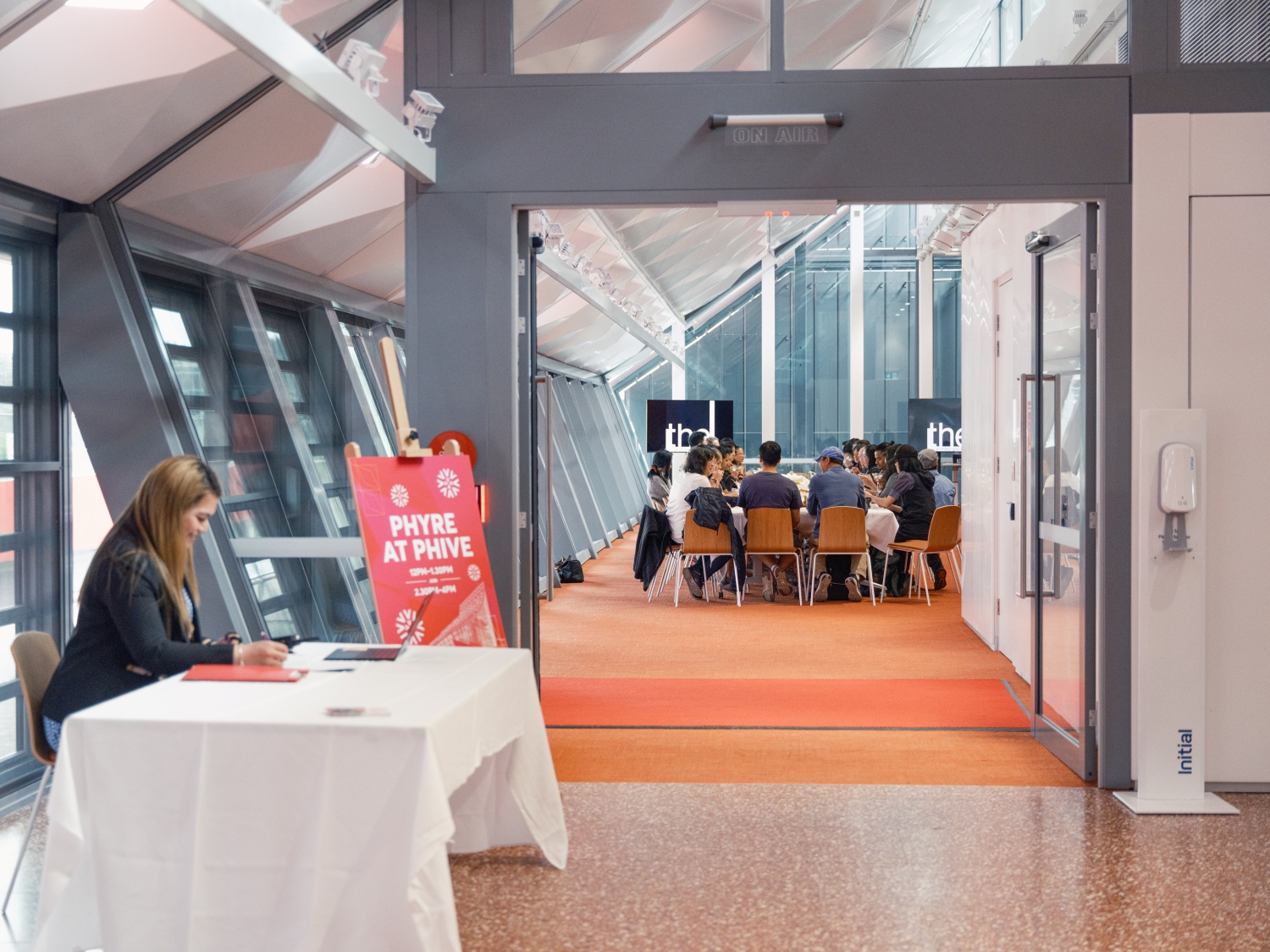 A woman sits at a table with a "Phyre at Phive" sign on the left. In the background, a group gathers for a meeting in a bright room. The scene is professional and welcoming.