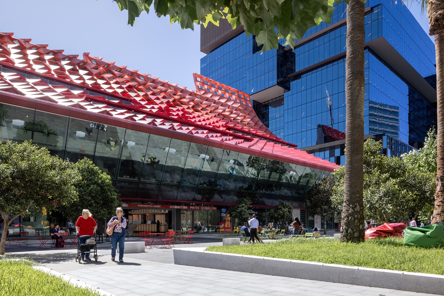 Modern architectural plaza with red angular roof and glass facade. People relax on chairs and benches amid greenery. A blue glass building contrasts behind.