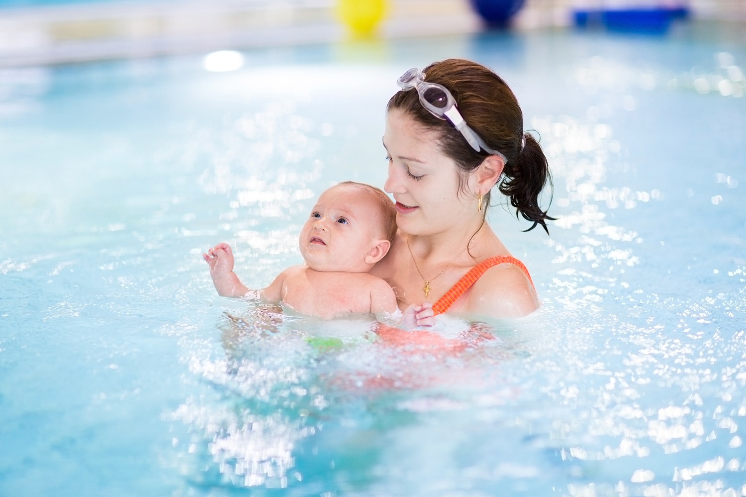 A woman in a red swimsuit is holding a baby in a swimming pool. She wears goggles on her head, smiling gently. The scene feels nurturing and joyful.