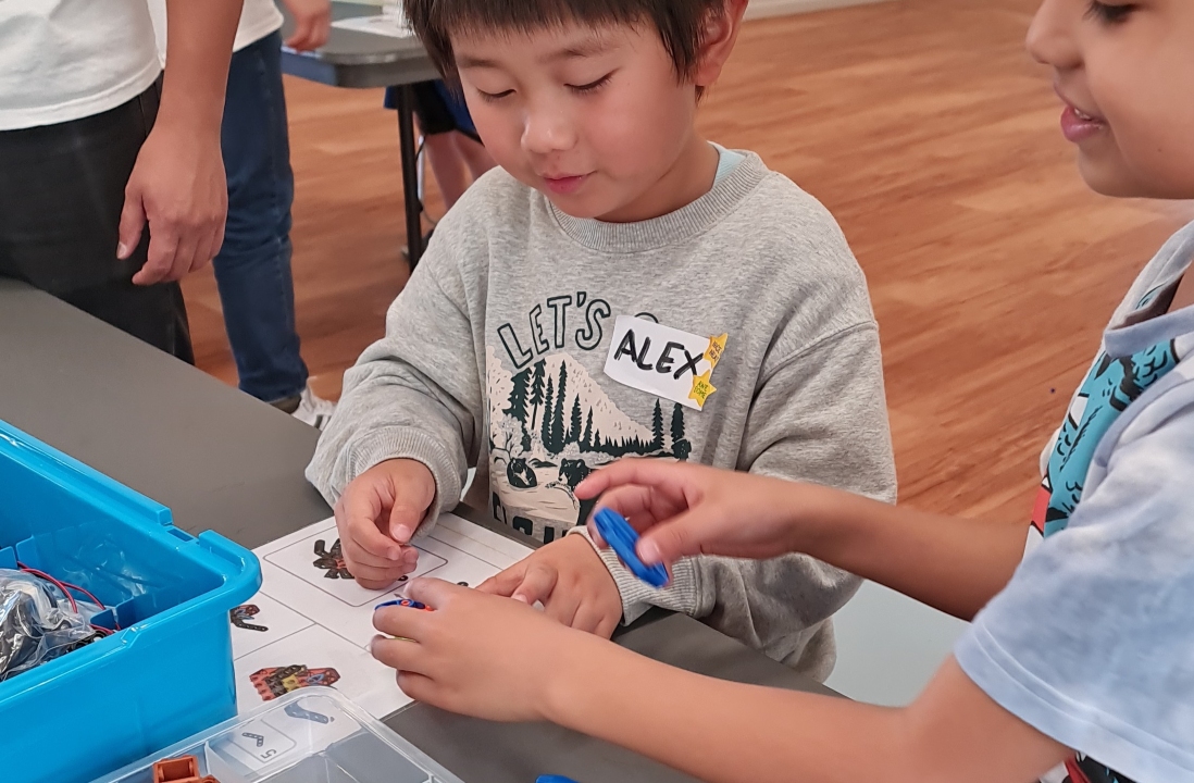 Two children sit at a table, focused and smiling, working on a robotic kit with a blue toolbox. The setting is a bright classroom.