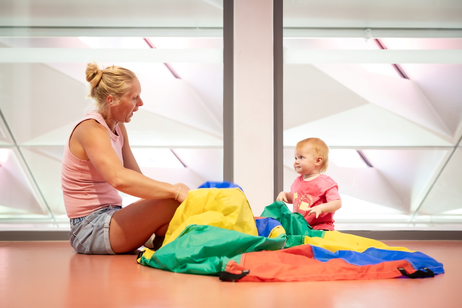 A woman and a baby play with a colorful parachute indoors. The woman sits cross-legged, smiling at the baby, who looks intrigued. Bright, lively atmosphere.