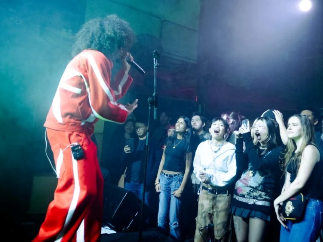 Performer in a red tracksuit sings passionately into mic on stage. Enthusiastic fans cheer and capture the moment, creating an energetic concert atmosphere.